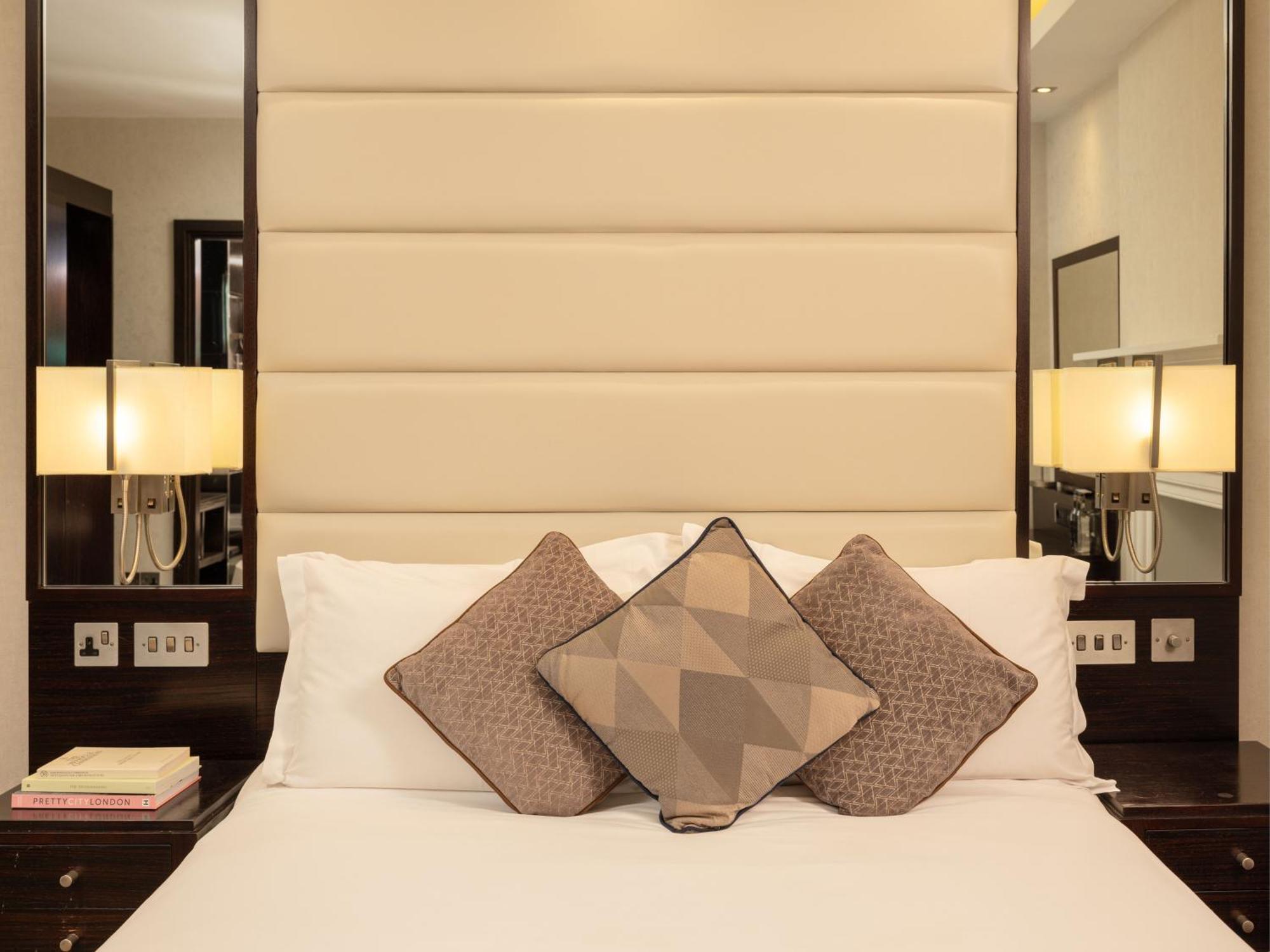 Hotel Montcalm Brewery Townhouse Londýn Exteriér fotografie The image shows a cozy bedroom setting. The bed features a headboard upholstered in a light beige fabric. It is adorned with three decorative pillows in varying patterns and colors, placed on white bedding. On either side of the bed, there are modern