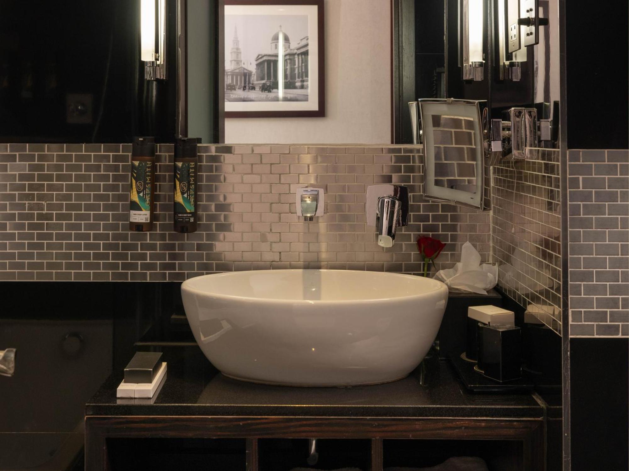Hotel Montcalm Brewery Townhouse Londýn Exteriér fotografie The photo shows a modern bathroom with a stylish design. It features a large, round white sink placed on a dark wooden vanity. The walls are adorned with a mix of gray and silver tile, creating a sleek and contemporary look. Above the sink, there is 