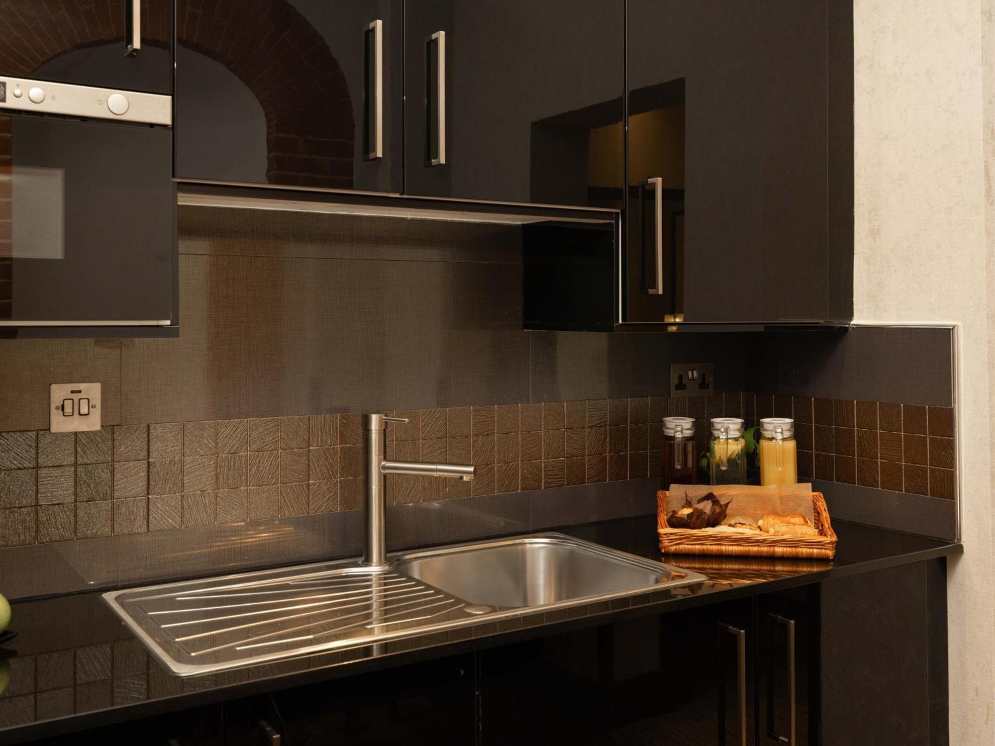 Hotel Montcalm Brewery Townhouse Londýn Exteriér fotografie The photo shows a modern kitchen with a sleek design. It features dark cabinetry with a glossy finish and stainless steel handles. There is a stainless steel sink with a simple faucet, and a countertop made of black material. Above the sink, there ar